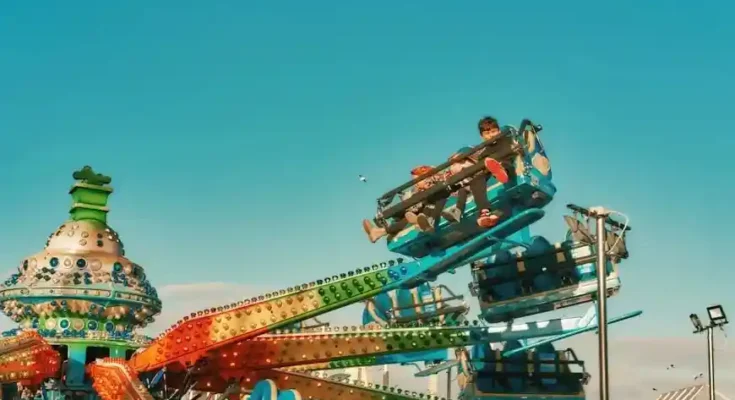 Two children enjoy a thrill ride at a carnival, suspended high against a bright blue sky filled with a few scattered clouds.