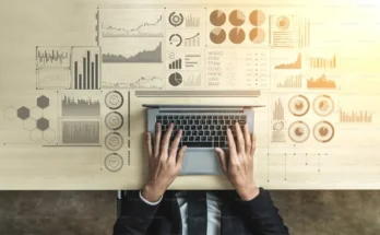 An overhead view of hands typing on a laptop surrounded by various analytical graphs and data visualizations on a wooden table.