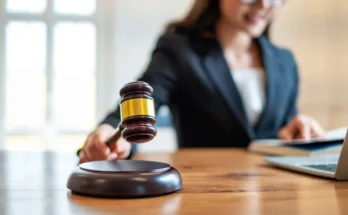 A judge in a black suit raises a gavel, with a laptop and documents on the desk, signaling the start of a court session.