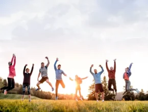 A joyful group of nine people jumping in the air, celebrating outdoors at sunset, surrounded by trees and green grass.