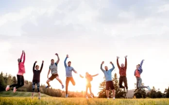 A joyful group of nine people jumping in the air, celebrating outdoors at sunset, surrounded by trees and green grass.