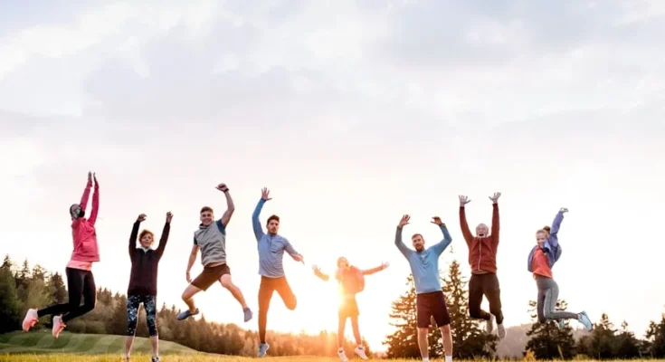 A joyful group of nine people jumping in the air, celebrating outdoors at sunset, surrounded by trees and green grass.
