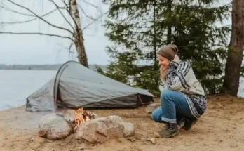 A person sits beside a campfire on a sandy shore, with a tent pitched nearby and trees framing a serene lake view.