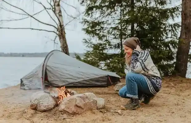 A person sits beside a campfire on a sandy shore, with a tent pitched nearby and trees framing a serene lake view.