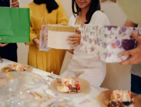 A group of friends at a celebration, holding colorful gift bags and boxes, with a table of desserts in front.