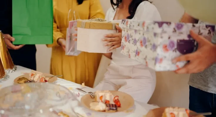 A group of friends at a celebration, holding colorful gift bags and boxes, with a table of desserts in front.